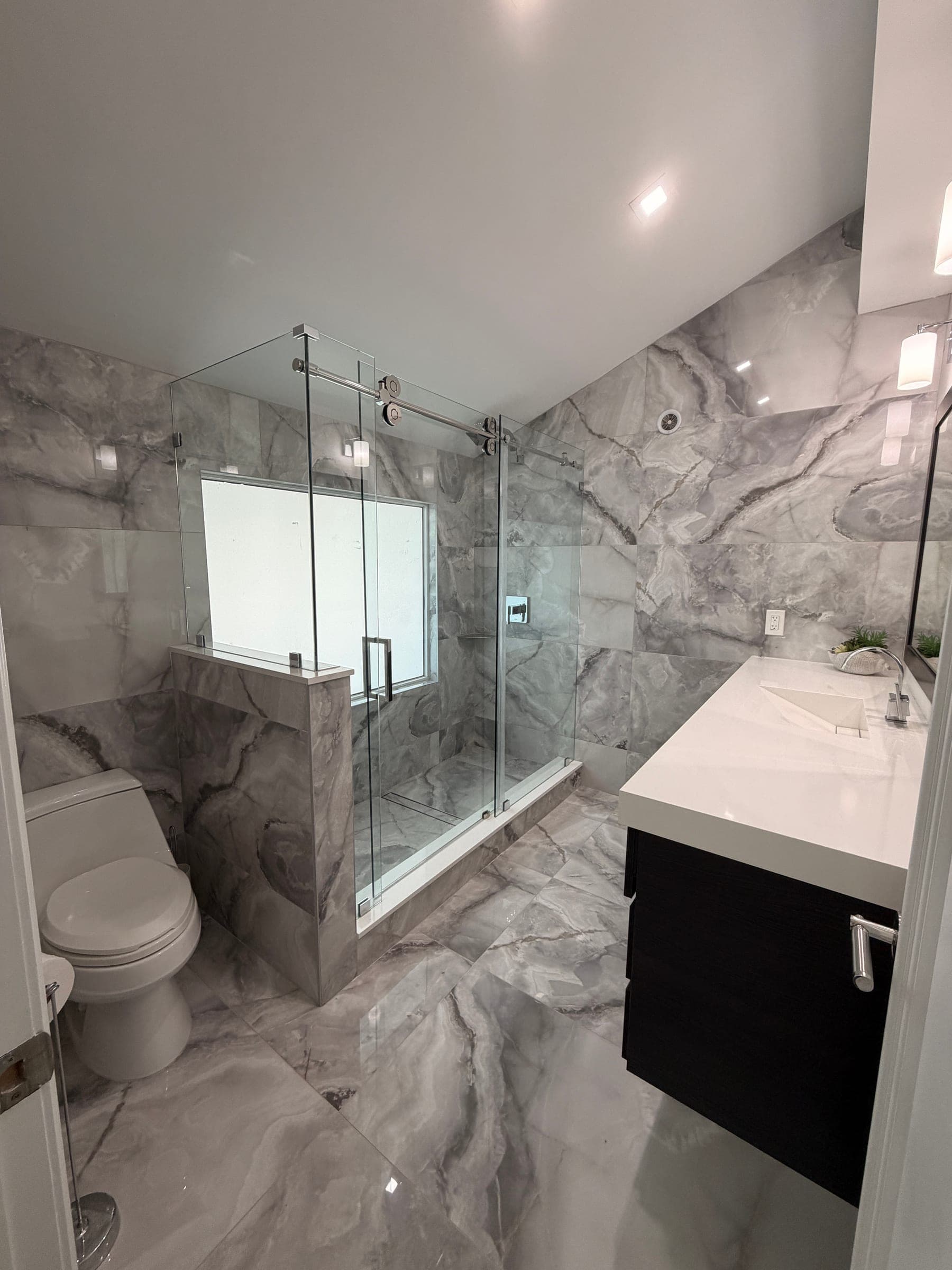 Remodeled bathroom with frameless glass shower enclosure, marble-look tile, and dark vanity with white countertop